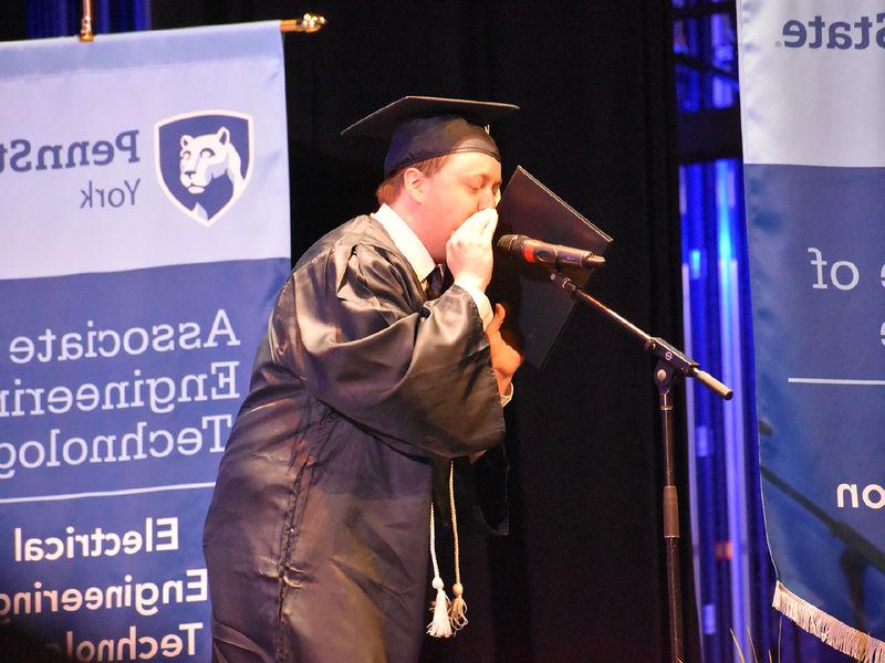 一名戴着毕业帽、身穿毕业服的男生站在凳子上对着麦克风讲话