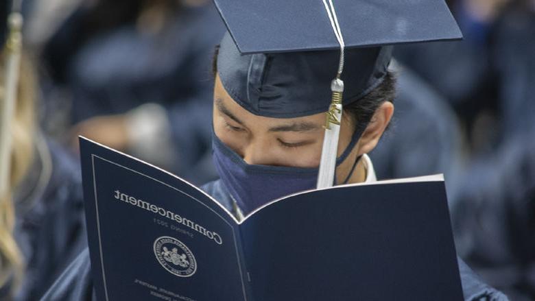graduate in cap, gown, and face mask reading program
