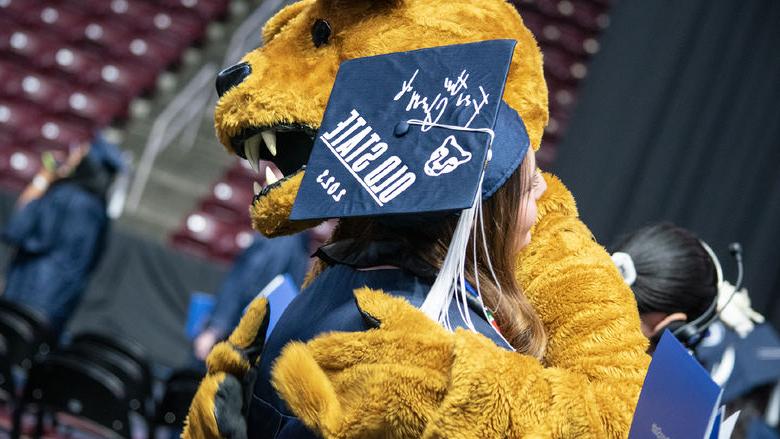 The Nittany Lion hugs a graduate in a cap that reads "For the Glory of Old State 2023"