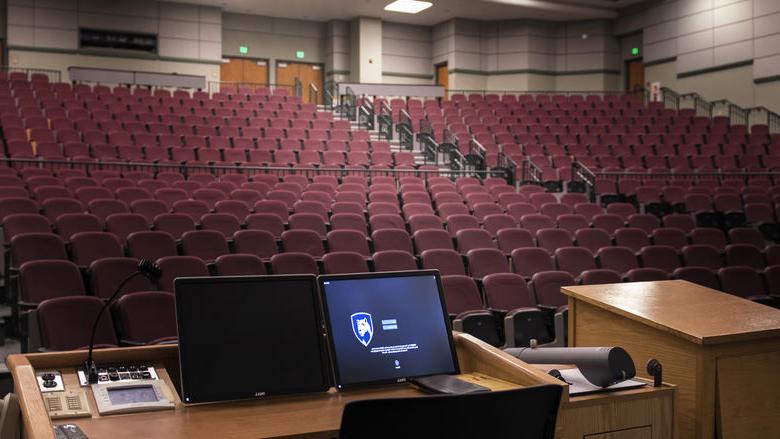 A computer screen is lit up at a podium overlooking an empty theater classroom.